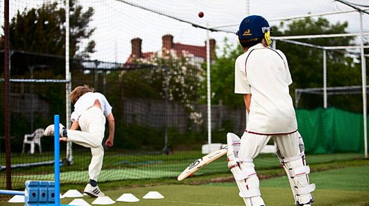 Young cricketers in full cricket attire practicing in a training net, wearing custom cricket jerseys, cricket whites, protective gear, and cricket pad covers, representing high-quality sports academy training.